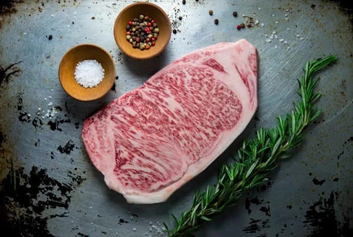 Raw A5 Wagyu steak showing intense marbling on a distressed metal surface with sea salt, peppercorns, and rosemary.