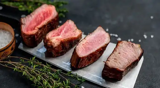 Four slices of cooked beef steak showing different levels of steak doneness from rare to well done lined up on a metal platter.