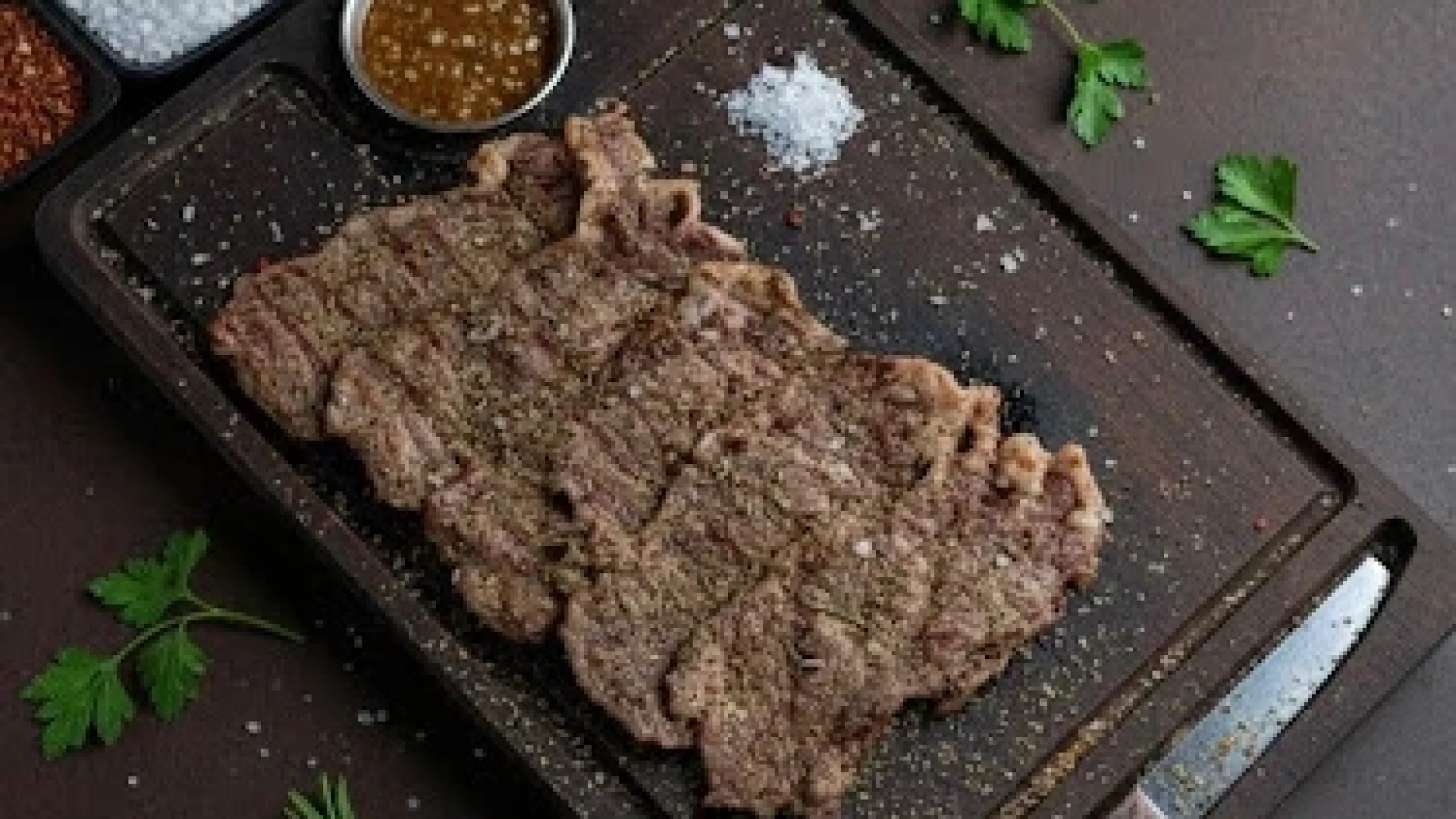 Seasoned grilled striploin steaks arranged on a dark wooden cutting board with coarse salt and fresh herbs.