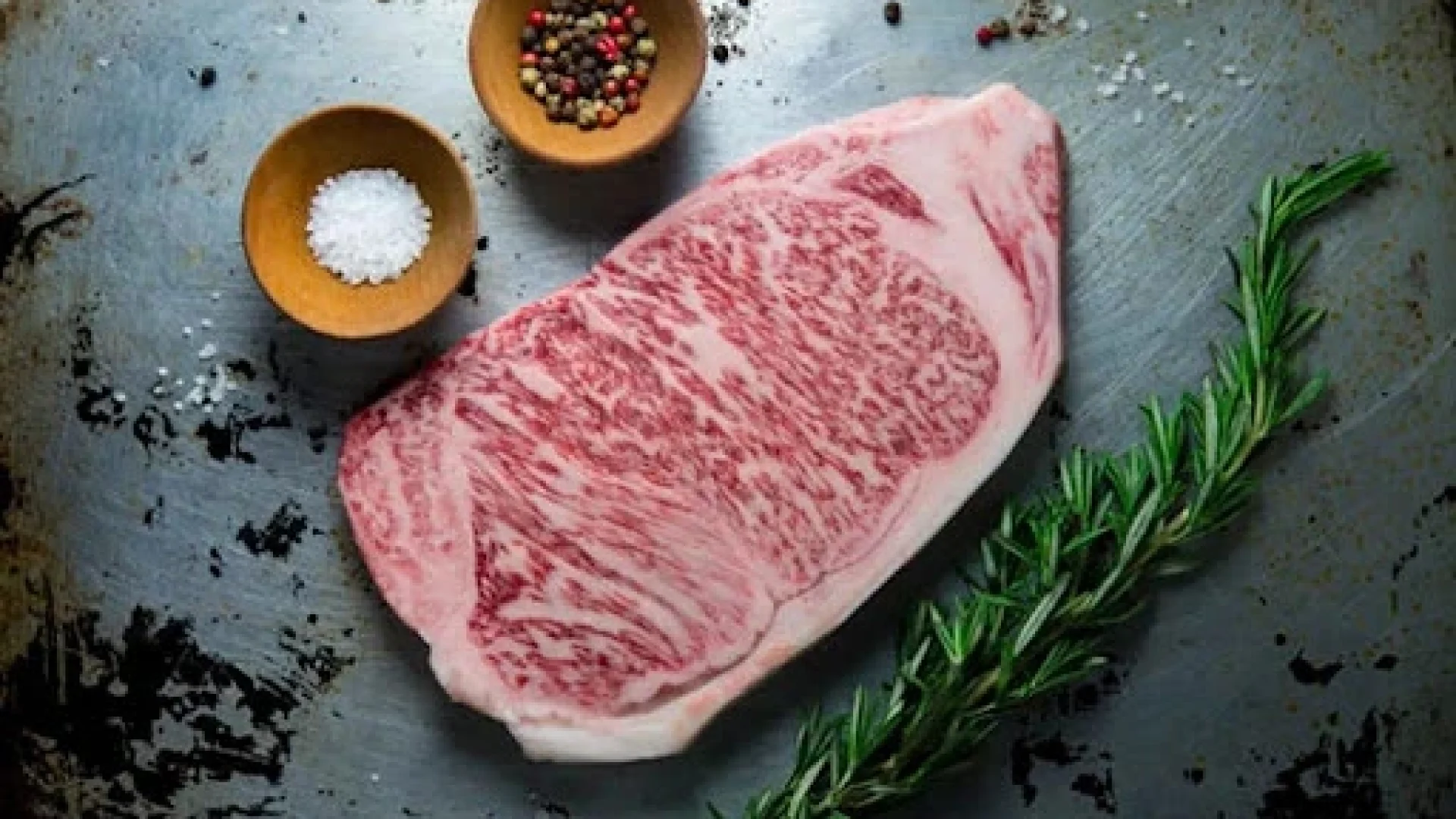 Raw A5 Wagyu steak showing intense marbling on a distressed metal surface with sea salt, peppercorns, and rosemary.