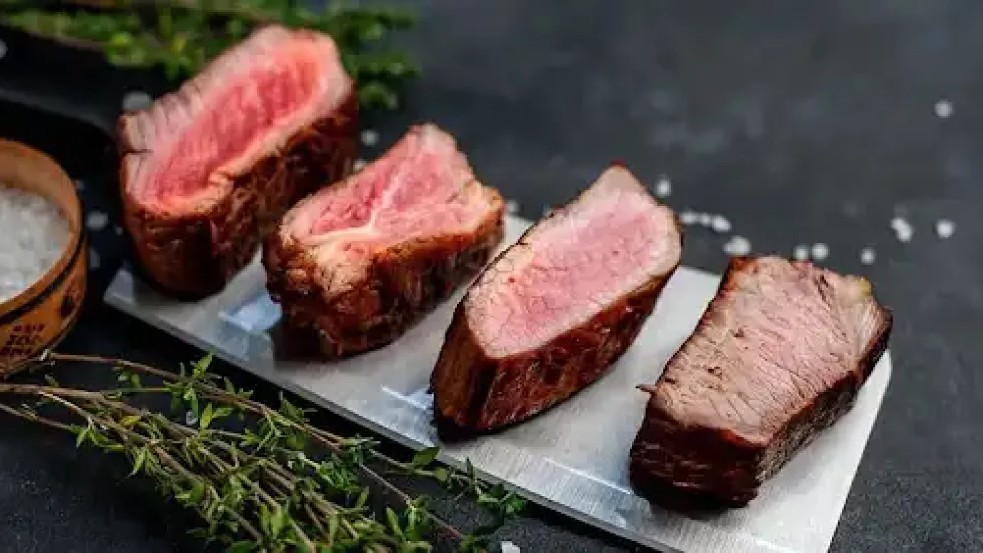 Four slices of cooked beef steak showing different levels of steak doneness from rare to well done lined up on a metal platter.