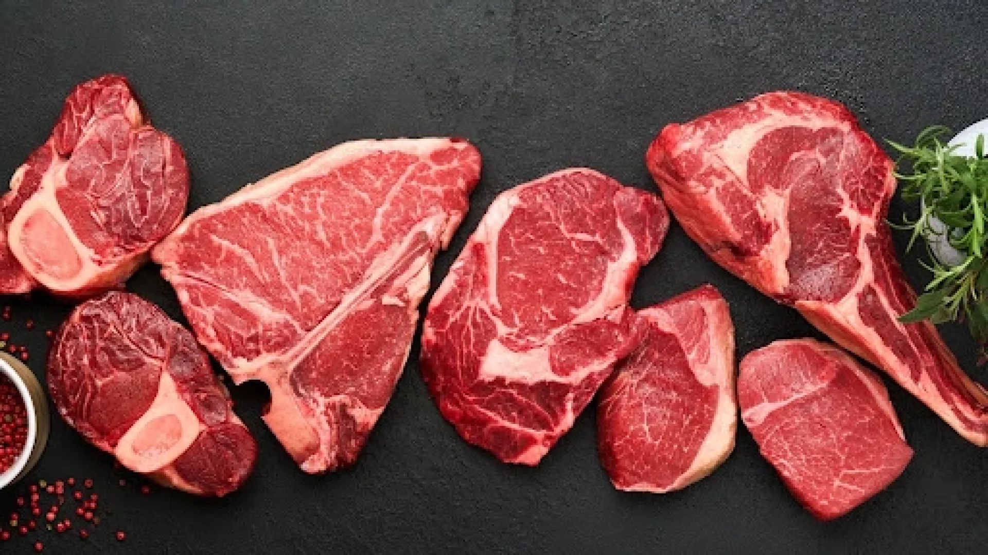 Assortment of raw steak cuts including bone-in ribeye, T-bone, sirloin, and filet arranged on a dark surface with peppercorns and fresh herbs