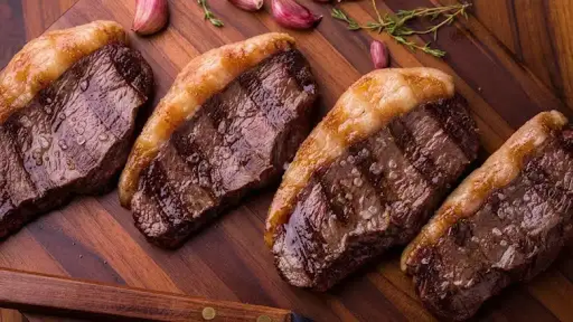 Grilled picanha steak slices with seared fat cap, coarse salt, garlic cloves, and fresh rosemary on a wooden board